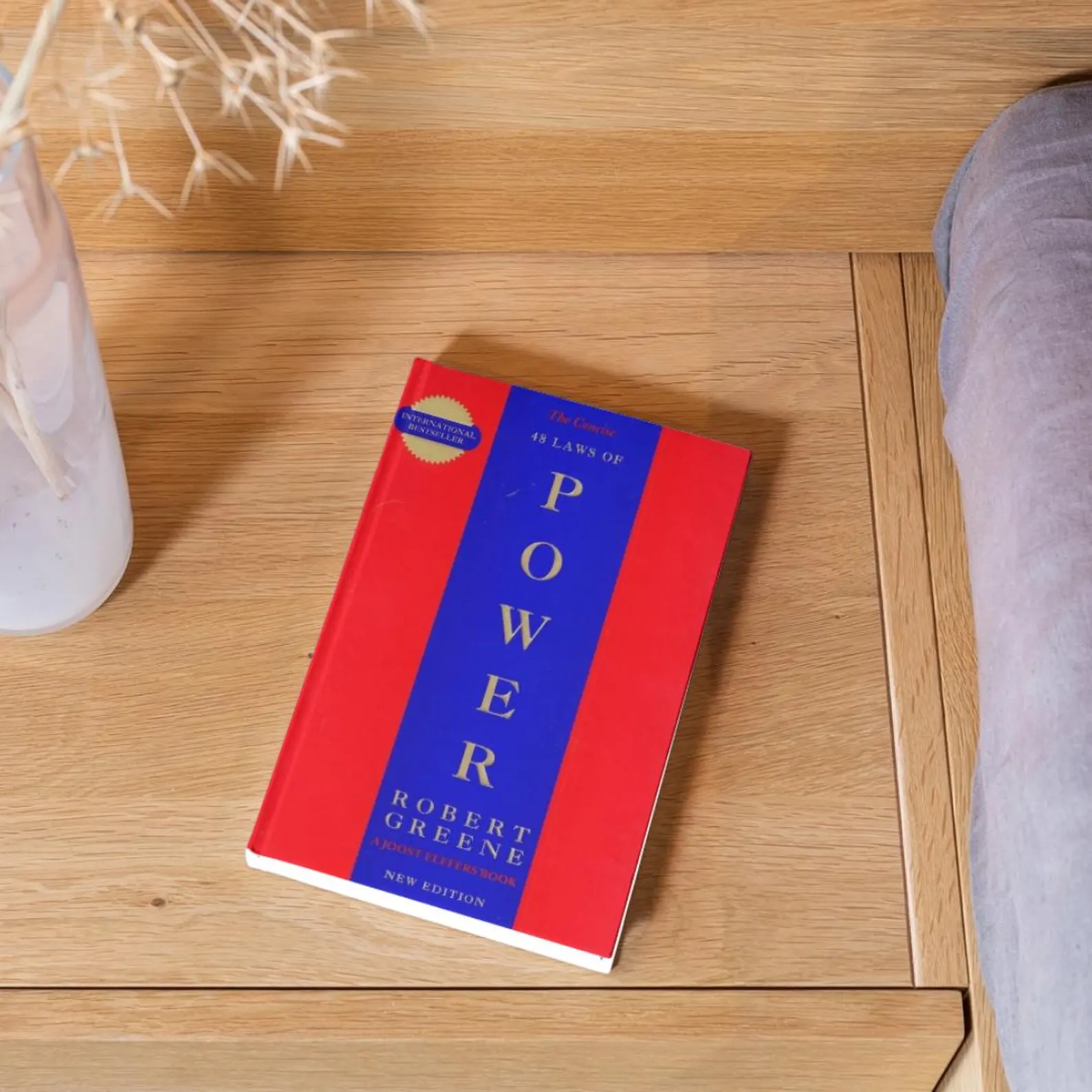 Le manuel rouge du pouvoir de Robert Greene Le livre "The 48 Laws of Power" de Robert Greene posé sur une table en bois clair à côté d’un vase blanc.